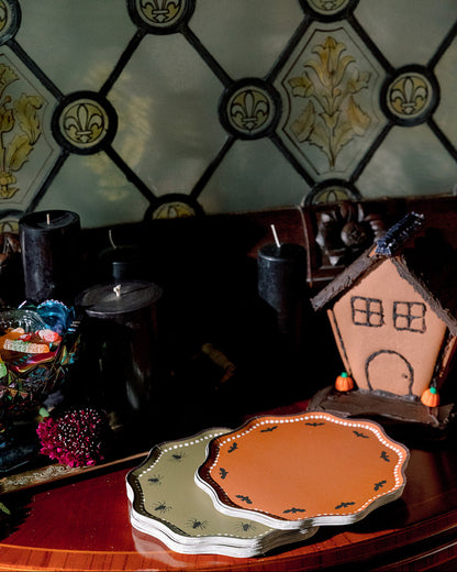 A styled tabletop scene with a small gingerbread house decoration, black candles, and two scalloped plates in orange and sage green in front of a stained-glass-style wall.