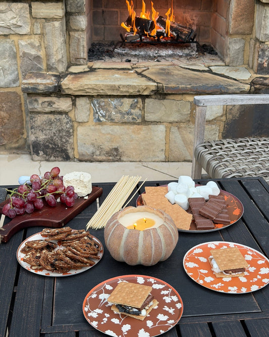 A cozy outdoor s'mores setup on a dark wooden table in front of a stone fireplace. Plates hold graham crackers, marshmallows, chocolate bars, pretzels, grapes, and a small pumpkin-shaped candle, with s'mores being assembled.
