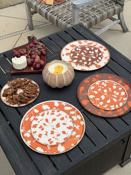 A set of orange and white autumn-themed plates with a maple leaf pattern, styled on a dark outdoor patio table with food and a candle.
