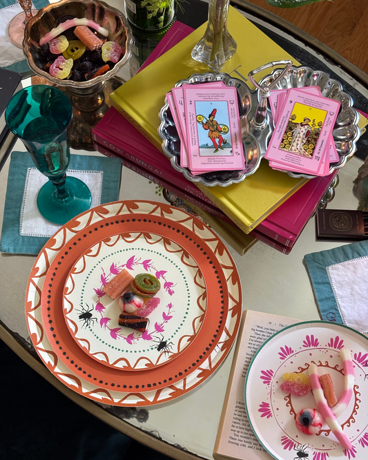 A styled tabletop scene with decorative plates, pink tarot cards, colorful books, and candy on a glass table. The most prominent sellable item appears to be the patterned plate in the foreground, shown in a real interior setting rather than as an isolated studio packshot, so this fits Lifestyle rather than Product/Packaging.