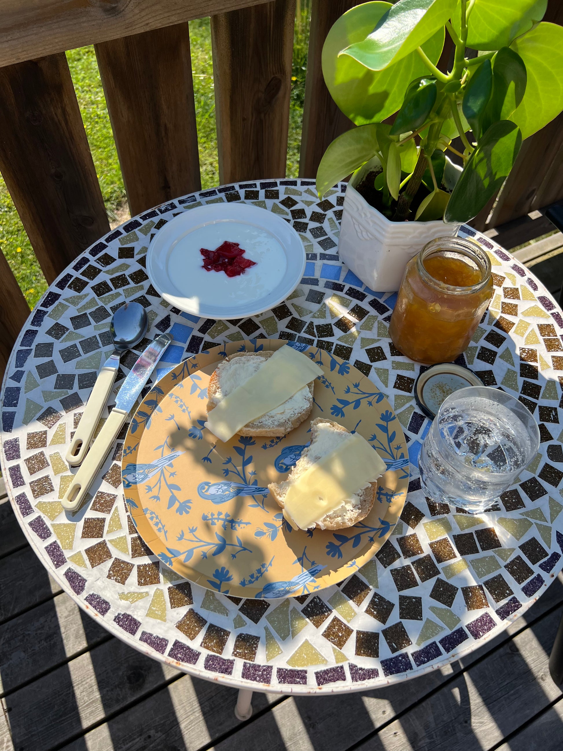 Mosaic table with plates of food, a jar of honey, and a glass on a wooden deck.