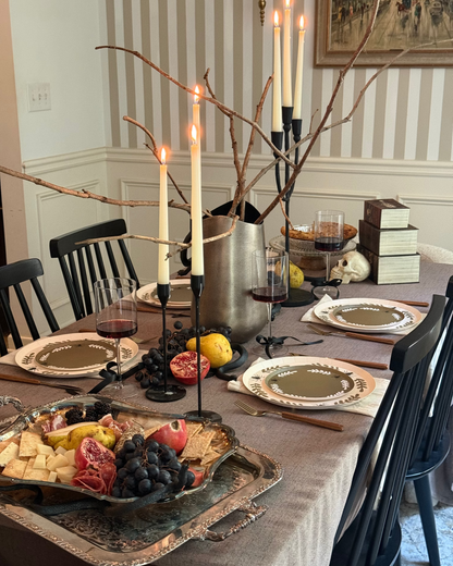 A styled dining table in a home setting, featuring a beige tablecloth, place settings with gold-rimmed plates, a charcuterie board with cheese, grapes, and cured meat, a silver bucket centerpiece with bare branches and lit taper candles, and a striped wall in the background.