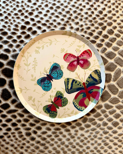 A round decorative plate  with a cream background, featuring a botanical pattern and four colorful illustrated butterflies in blue, green, pink, and red, placed on a textured leopard-print surface.