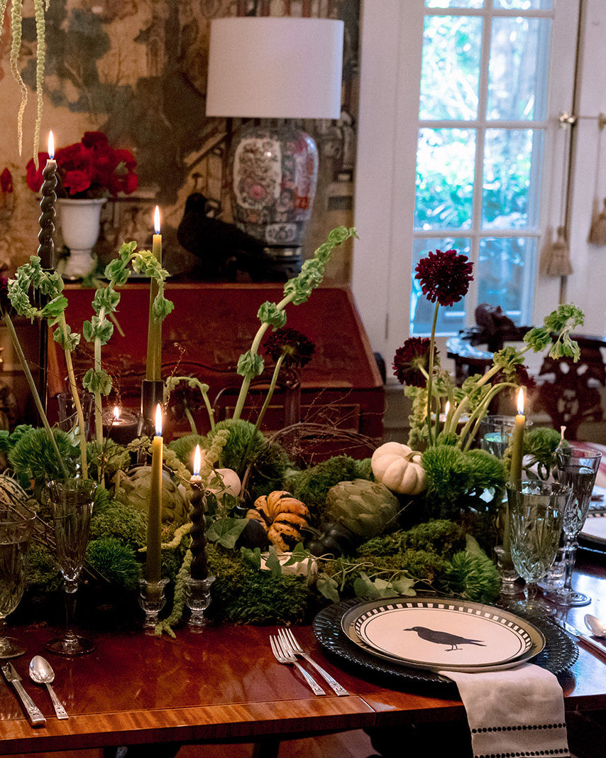 An elaborate autumn-themed table centerpiece with greenery, candles, pumpkins, and place settings in a decorated dining room.
