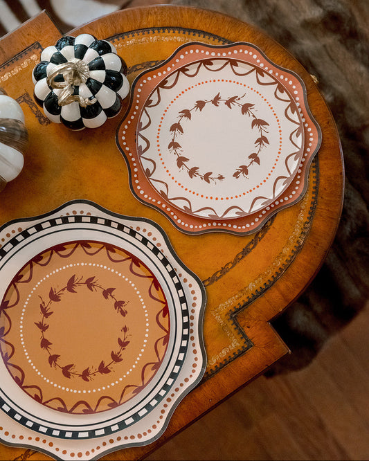 Two scalloped plates with autumn-themed orange, brown, and cream patterns, styled on a wooden side table next to a black and white checkered pumpkin.