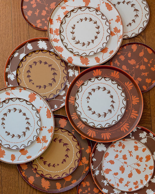 A collection of plates with autumn-themed patterns, including orange and brown leaves, floral motifs, and wreaths on a white background, arranged on a wooden table.