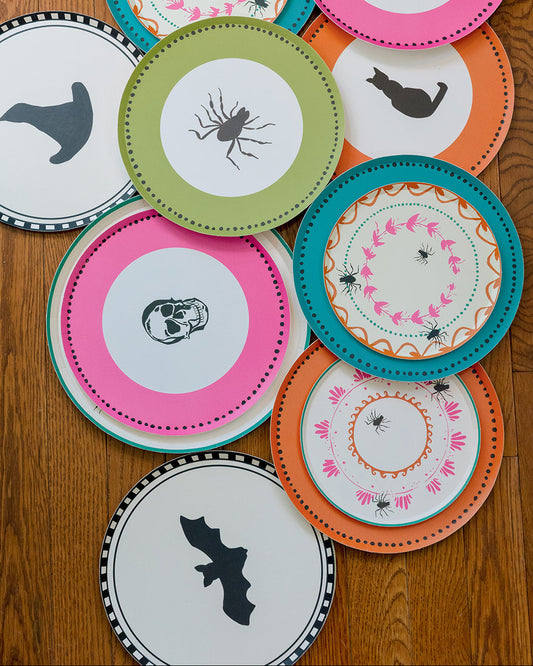 A collection of colorful Halloween-themed paper plates arranged on a wooden tabletop. The plates feature various spooky designs including skulls, bats, spiders, and a witch's hat, with vibrant pink, teal, orange, green, and white colors.
