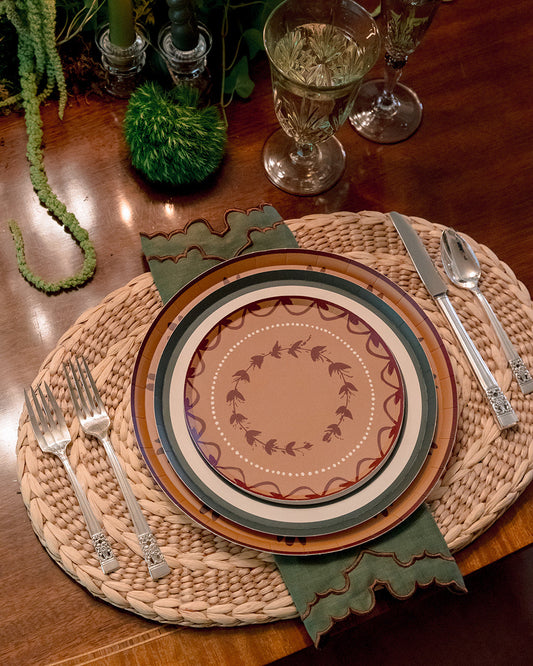 A styled table setting featuring a woven round placemat, a layered ceramic plate with a botanical wreath pattern, green scalloped napkins, silver flatware, and candle holders on a dark wood table.