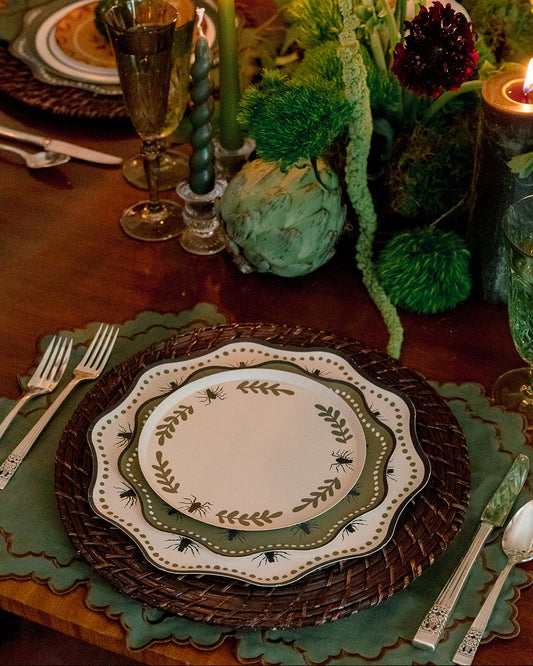A styled table setting with layered plates featuring a green leaf border, a dark brown woven charger, and green placemats on a wooden table, accented by lit candles and a green floral centerpiece.
