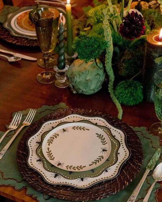 A styled table setting with layered plates featuring a green leaf border, a dark brown woven charger, and green placemats on a wooden table, accented by lit candles and a green floral centerpiece.