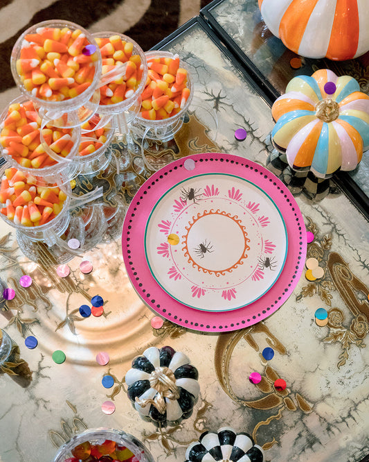 A styled Halloween table setting featuring a pink and white patterned plate, clear cups filled with candy corn, small decorative pumpkins, and scattered confetti on a reflective marble-patterned tabletop.