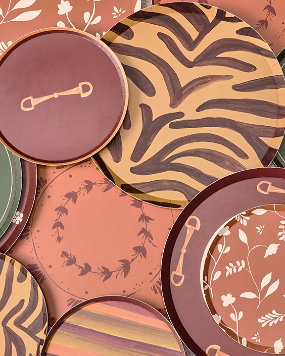 A close-up arrangement of decorative round plates with coordinated patterns in terracotta, rust, brown, beige, and muted green. Designs include zebra-like brushstroke motifs, floral vine borders, wreath details, and small gold-toned accents.