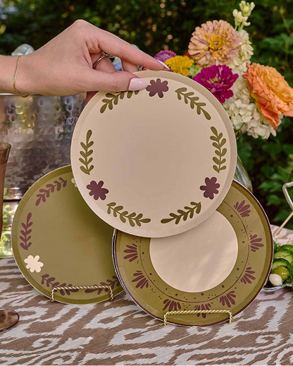 Three decorative plates with a floral and leaf pattern are displayed outdoors on a table. A hand is holding the top plate, which is beige with olive green leaves and purple flowers.