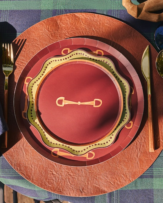 A styled table setting featuring a rustic place setting with a navy blue napkin, gold-toned cutlery, and a layered plate set in terracotta, rust, and olive green on a green plaid tablecloth.
