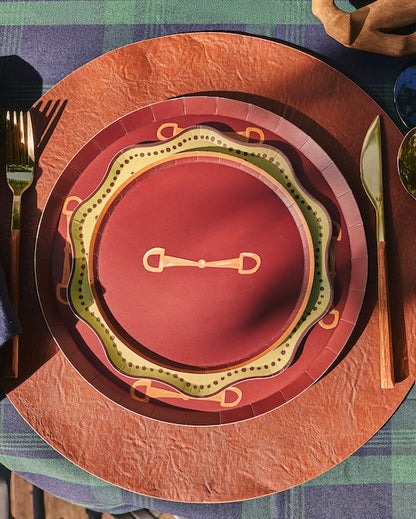 A styled table setting featuring a rustic place setting with a navy blue napkin, gold-toned cutlery, and a layered plate set in terracotta, rust, and olive green on a green plaid tablecloth.