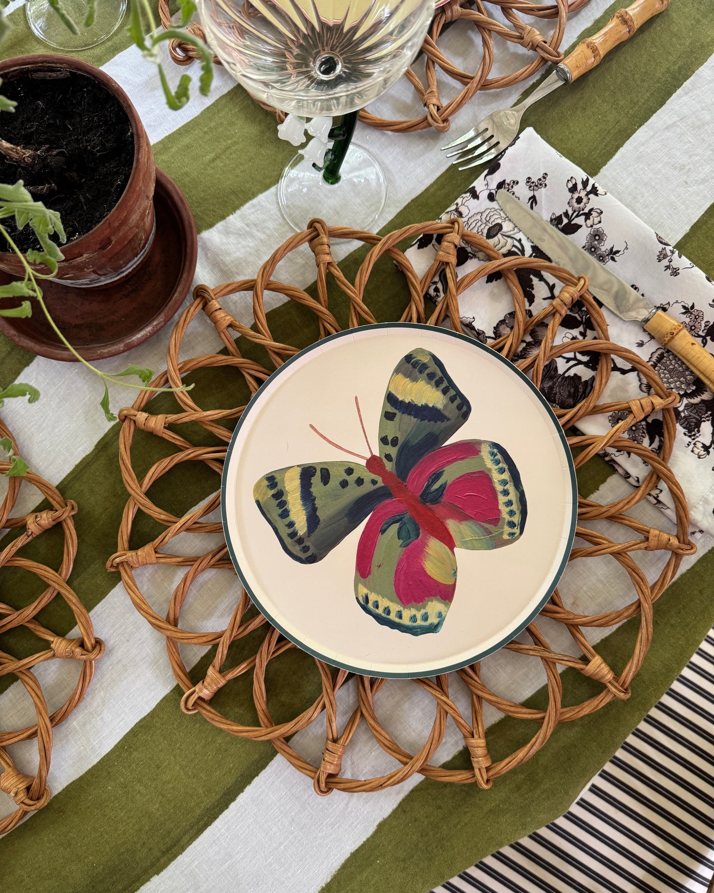 A styled tabletop setting featuring a round plate with a colorful butterfly illustration, placed on a woven rattan placemat over a green and white striped tablecloth.