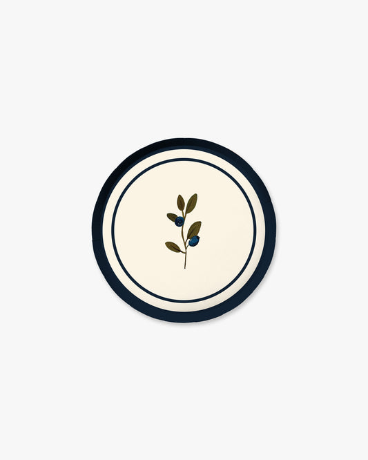 A round decorative plate featuring a cream center with a small botanical illustration of a leafy olive branch, framed by a dark navy blue border and a thin white ring.
