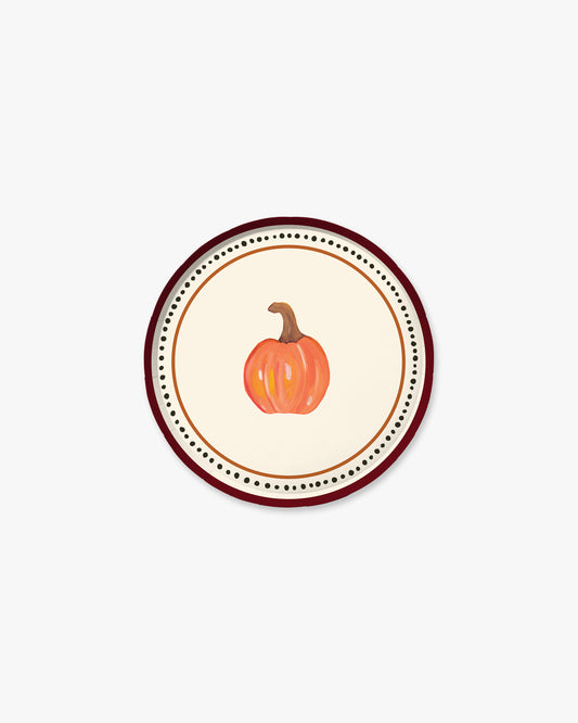 A round decorative plate featuring a central illustration of a small orange pumpkin with a brown stem, surrounded by concentric rings of black dots and a dark red outer border on a light cream background.