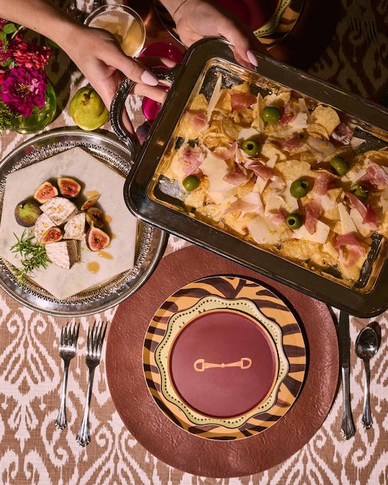An overhead view of a styled dining table with patterned brown and gold plates, silver cutlery, a tray of baked pasta topped with ham and green olives, a plated salad, and hands reaching in from the top holding a glass.