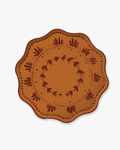 A round decorative plate with a scalloped edge, featuring a warm terracotta orange-brown center and a dark brown printed floral wreath pattern, shown on a light gray background.