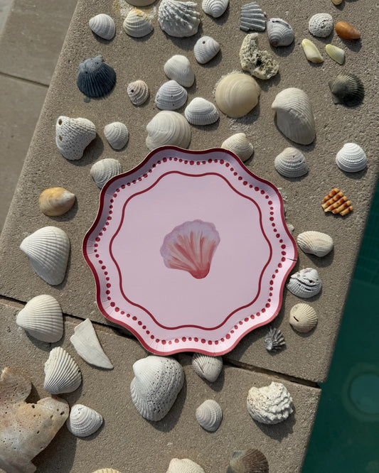 A pink decorative plate with a shell motif in the center on a cement background with shells scattered.