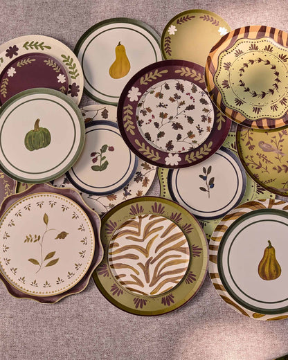 A collection of decorative plates with autumn-themed patterns, including pumpkins, gourds, leaves, and floral motifs in earthy tones of green, brown, purple, and beige, arranged on a textured gray fabric background.