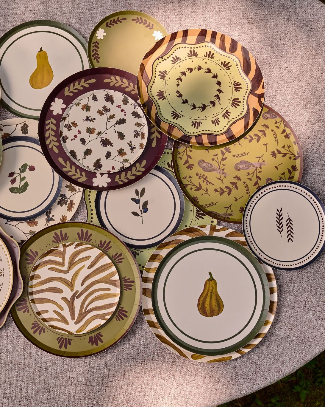 A collection of decorative plates with autumn-themed patterns, including pumpkins, pears, leaves, and floral motifs, arranged on a round gray table.