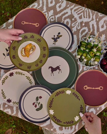 A styled outdoor dining scene showing a collection of decorative plates with horse and botanical designs, arranged on a patterned tablecloth next to a wicker chair on grass.