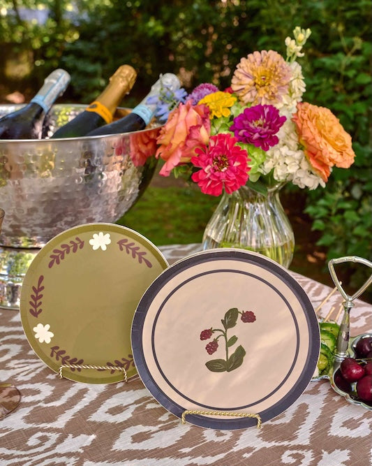 Two decorative plates displayed on a patterned tablecloth outdoors, with a floral arrangement and a metal ice bucket in the background.