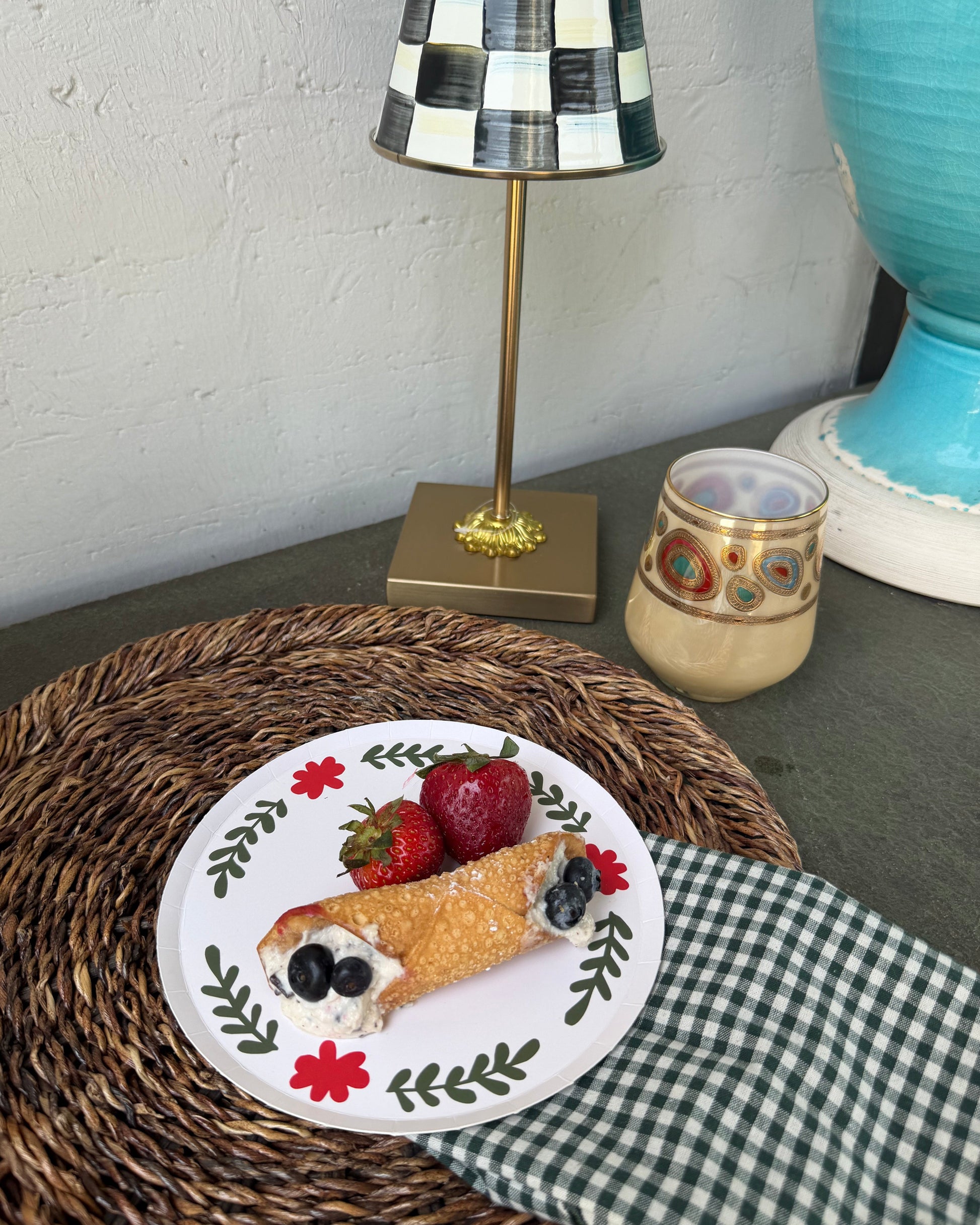 A plated dessert with strawberries and blueberries sits on a woven placemat next to a green-and-white checkered napkin, with a decorative lamp and ceramic vessel in the background.