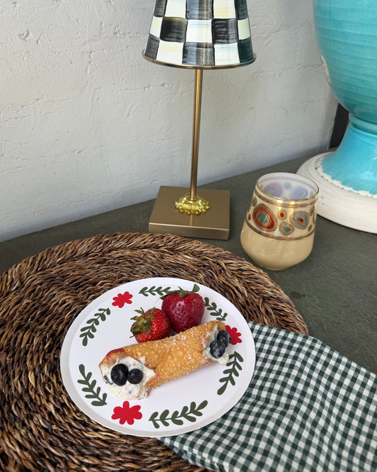 A plated dessert with strawberries and blueberries sits on a woven placemat next to a green-and-white checkered napkin, with a decorative lamp and ceramic vessel in the background.
