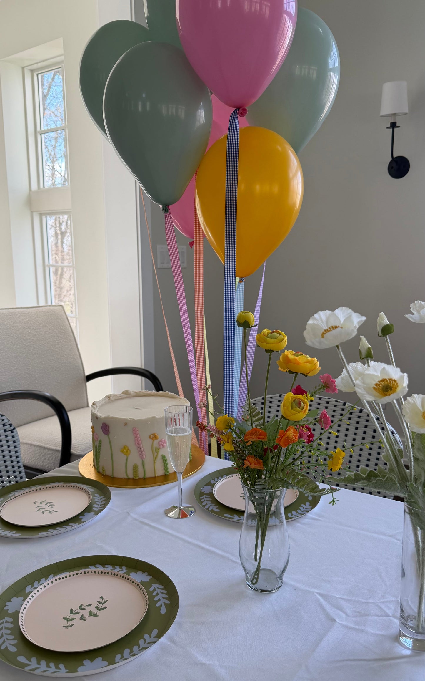 Dining table set with plates, a cake, and balloons in a bright room.