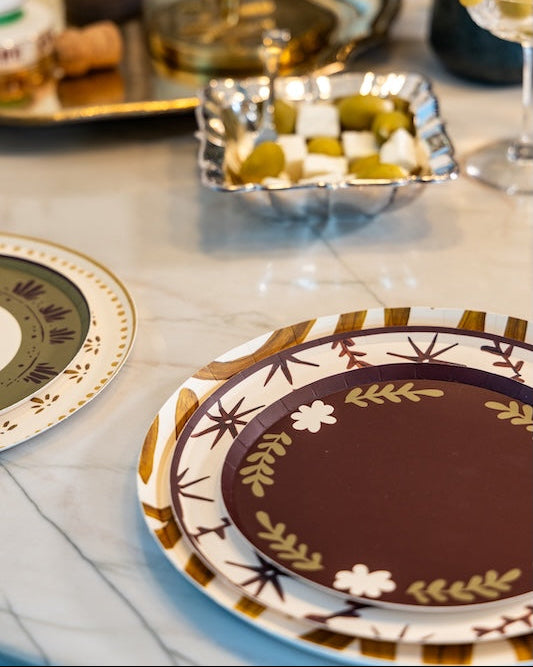 A close-up of decorative dinnerware on a marble table, featuring a round plate with a dark brown center and a gold and white floral border, stacked on another matching plate.