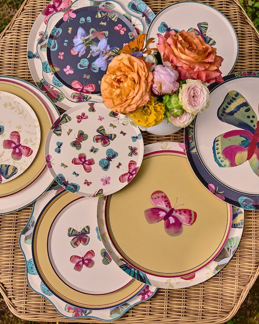 A collection of decorative ceramic plates with colorful butterfly and floral patterns, arranged on a woven wicker table with a small bouquet of orange and pink roses, set against a background of green grass.