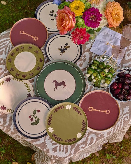 A styled outdoor dining scene showing a collection of decorative plates with horse and botanical designs, arranged on a patterned tablecloth next to a wicker chair on grass.