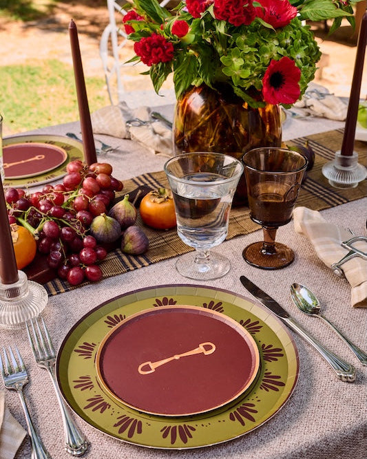 A styled outdoor table setting with a place setting featuring a deep red plate with a gold key motif, surrounded by a green and gold patterned charger plate. The table is decorated with a floral centerpiece, red candles, glasses, and fresh fruit.