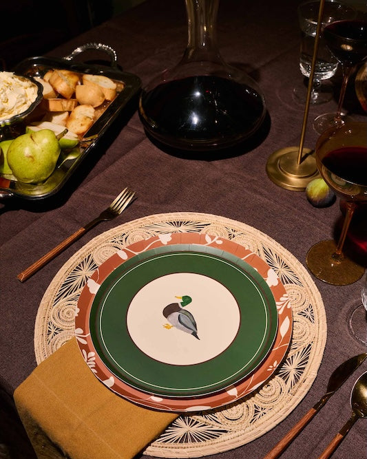 A styled dinner table setting featuring a place setting with a green and white plate printed with a mallard duck, layered over a brown and beige patterned charger plate and a woven placemat. A folded mustard yellow napkin, wooden-handled cutlery, and a dark brown tablecloth complete the rustic table arrangement.