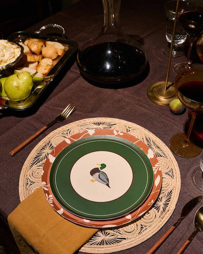 A styled dinner table setting featuring a place setting with a green and white plate printed with a mallard duck, layered over a brown and beige patterned charger plate and a woven placemat. A folded mustard yellow napkin, wooden-handled cutlery, and a dark brown tablecloth complete the rustic table arrangement.