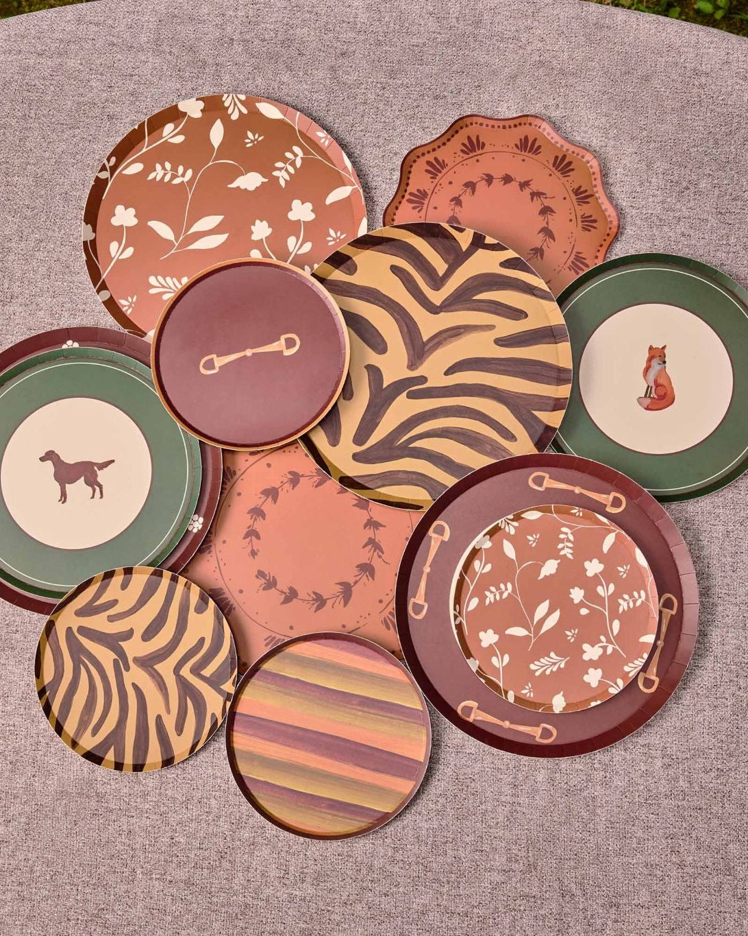 A group of decorative plates in earthy terracotta, brown, green, and beige tones, arranged on a round gray surface outdoors.