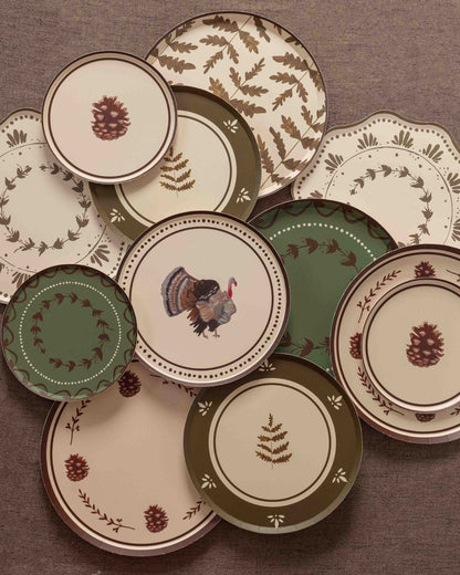 Overhead arrangement of fall-themed paper plates in cream, olive green, and burgundy featuring pinecone illustrations, botanical sprigs, fern patterns, floral wreaths, and a turkey motif on a textured brown backdrop.