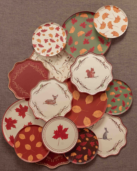 Overhead arrangement of autumn-themed decorative paper plates in cream, rust, burgundy, and sage green, featuring colorful fall leaves, maple leaf prints, and woodland animal illustrations of a deer, squirrel, and rabbit on a textured brown backdrop.