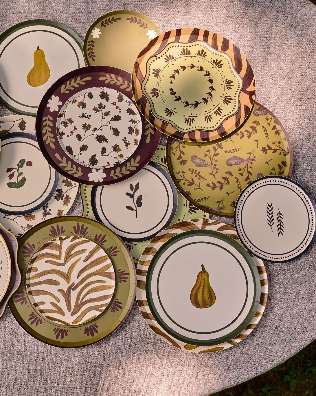 A collection of decorative plates with autumn-themed patterns, including pumpkins, gourds, leaves, and stripes, arranged on a light gray fabric-covered round table.