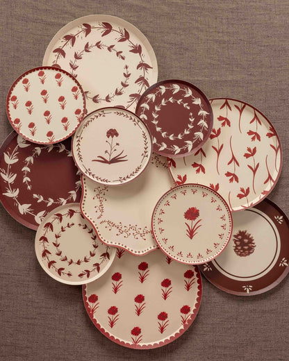 Overhead arrangement of decorative paper plates in cream, burgundy, and rust floral patterns, featuring botanical wreaths, delicate flower illustrations, and scalloped borders on a textured brown backdrop.