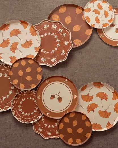 Overhead arrangement of autumn-themed decorative paper plates in cream, terracotta, and rust, featuring ginkgo leaves, delicate botanical wreaths, acorns, and scattered leaf patterns on a textured brown backdrop.