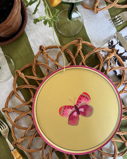 A round green plate with a pink butterfly illustration and pink rim, styled on a woven wicker placemat over a green and white tablecloth with cutlery and glassware.