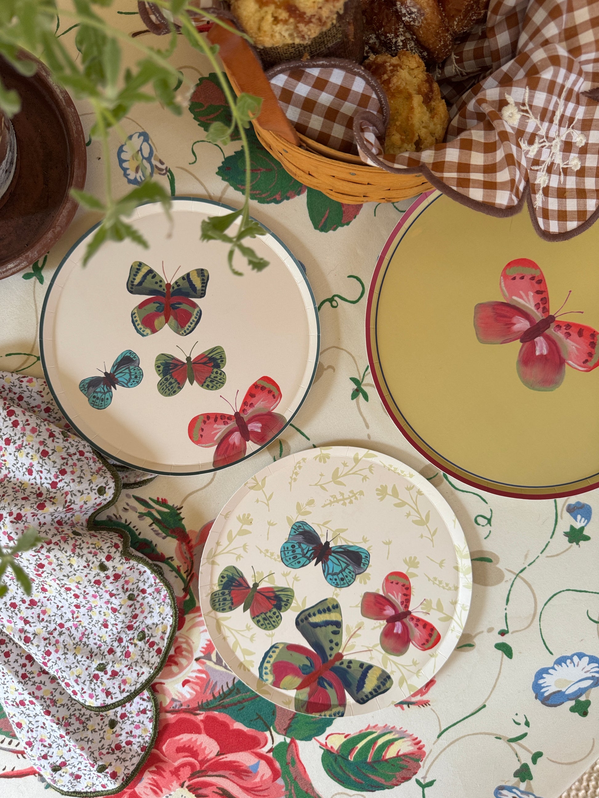 Three decorative plates with colorful butterfly and floral prints are arranged on a matching floral tablecloth, alongside a basket of muffins, a gingham napkin, and floral oven mitts.