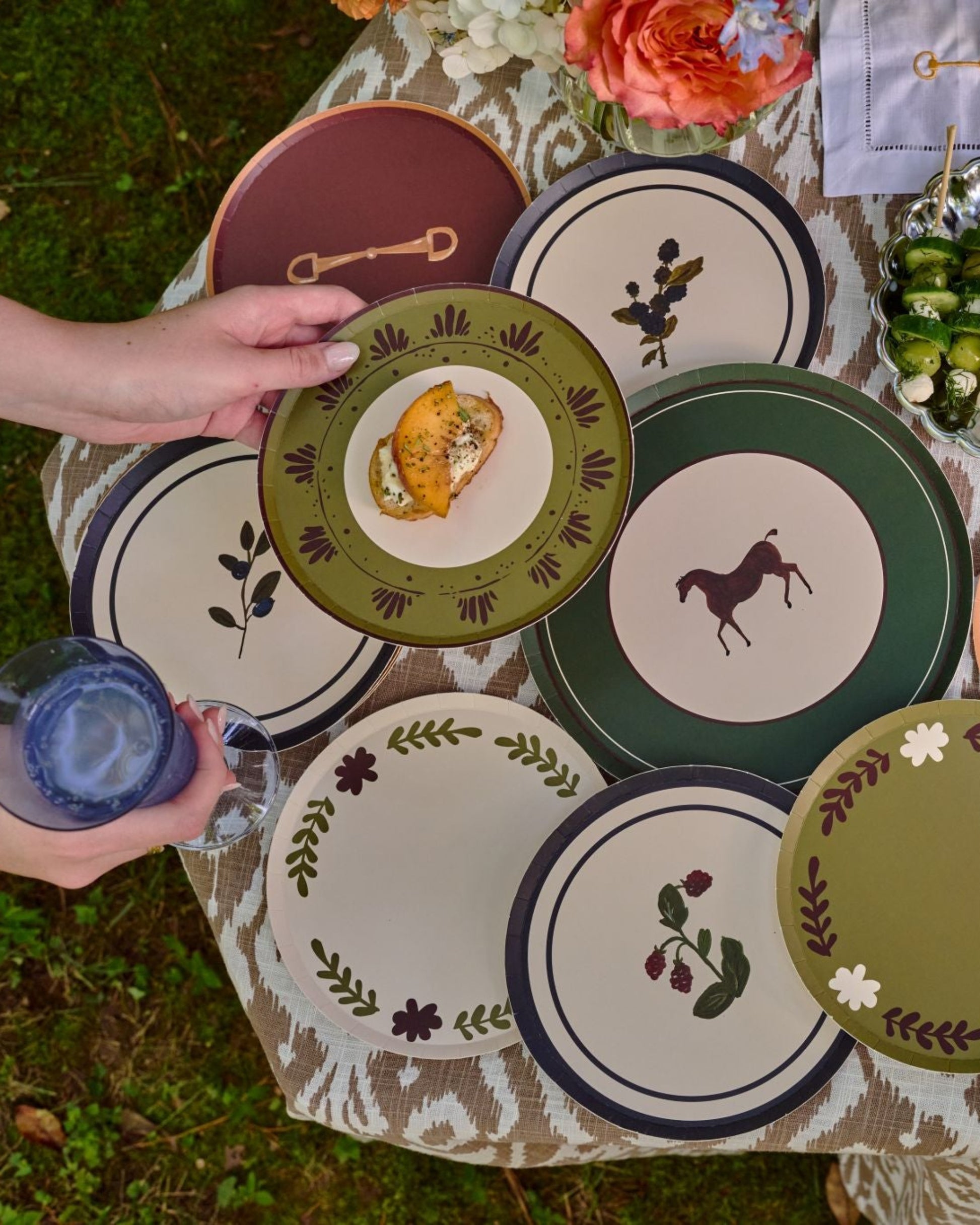A top-down outdoor view of a table covered with multiple decorative plates in green, cream, and burgundy, with hands holding plates. Bowls of fruit and a wicker basket are visible on a grassy lawn.