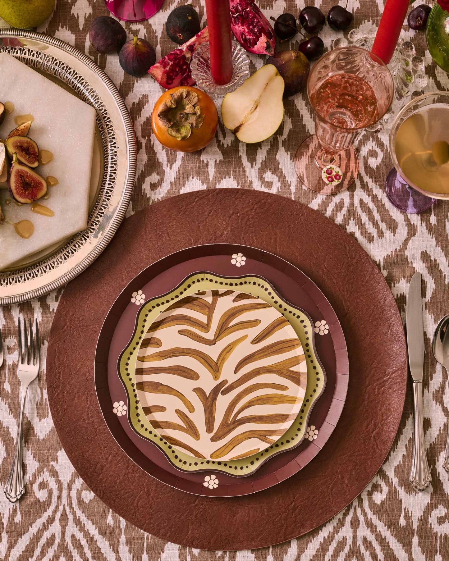 An overhead styled table setting featuring a round brown leather charger plate layered with a brown and black patterned plate, topped with a deep red center plate. The table is decorated with red taper candles, glasses, fresh figs, sliced apples, and a silver platter holding fig and white bean wraps on a patterned tablecloth.