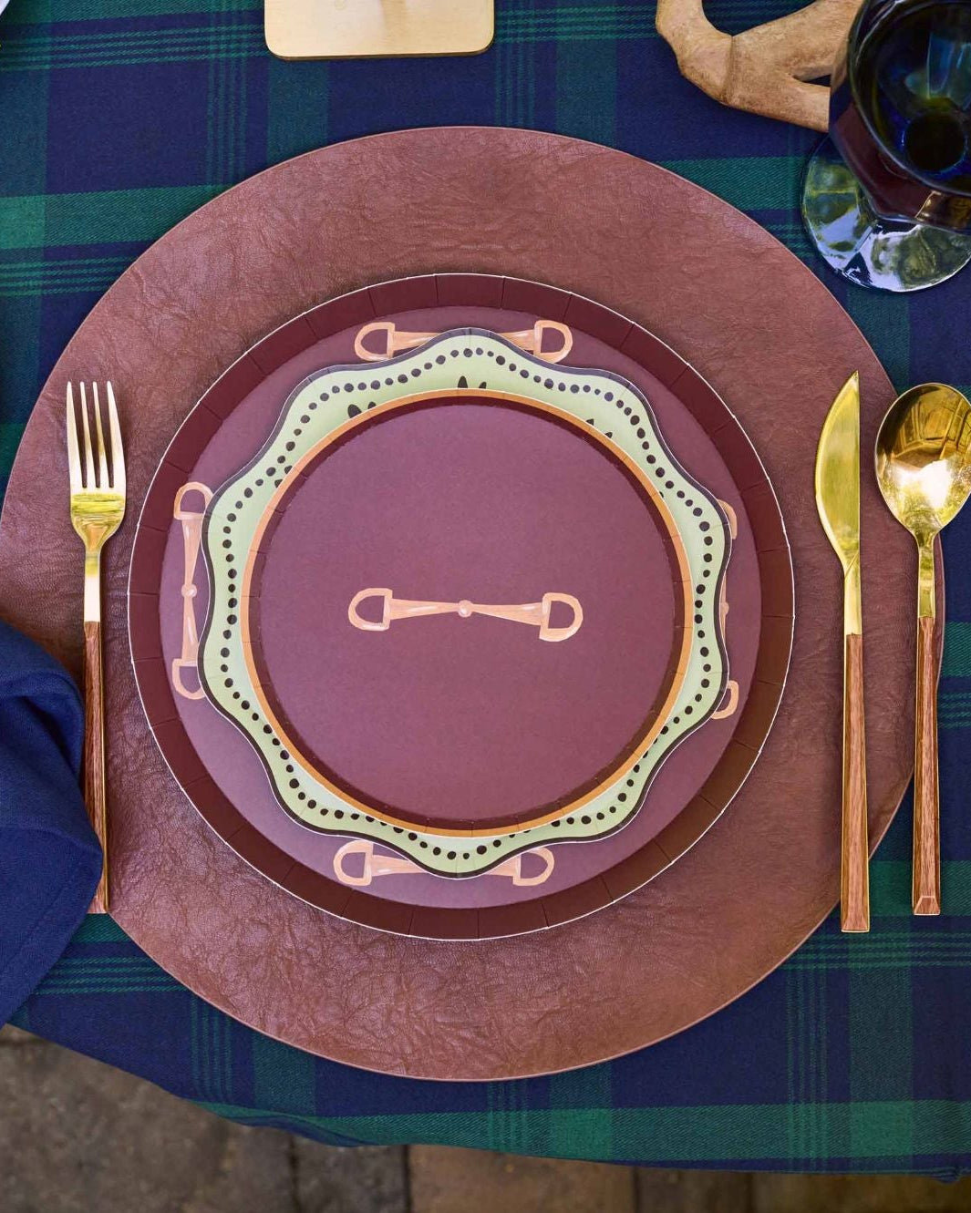 A styled table setting featuring a rustic place setting with a navy blue napkin, gold-toned cutlery, and a layered plate set in terracotta, rust, and olive green on a green plaid tablecloth.