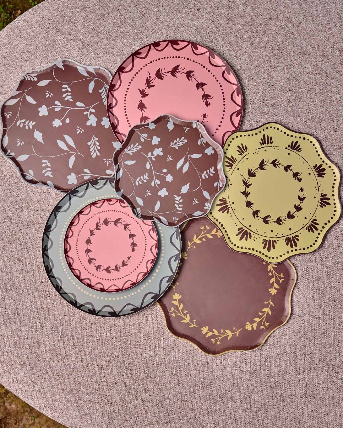 Decorative plates some with scalloped edges are arranged in a cluster on a round table. The plates feature various patterns and colors including pink, brown, yellow, and gray, with some showing floral motifs and others displaying geometric or lace-like borders.